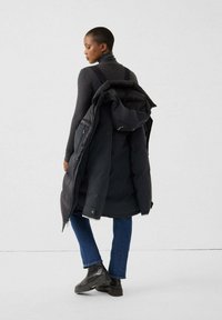 Vrouw in een donkere coltrui, blauwe jeans, zwarte laarzen, met een zwarte pufferjack als rugzak gedragen, staande tegen een effen lichte achtergrond.