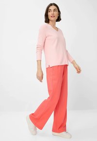 Rosa gerippter Langarmpullover mit V-Ausschnitt und seitlichen Schlitzen, kombiniert mit korallfarbenen weiten Hosen und weißen Sneakers vor einem einfarbigen Hintergrund.