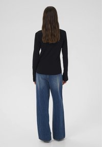 Žena s dlhými hnedými vlasmi oblečená v čiernej blúzke s dlhými rukávmi a širokých modrých džínsoch, stojaca chrbtom k jednofarebnému pozadiu.