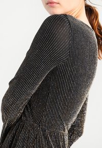 Robe noire avec des rayures verticales dorées, présentant une finition texturée et un corsage ajusté. Les manches sont transparentes, apportant un contraste au design.