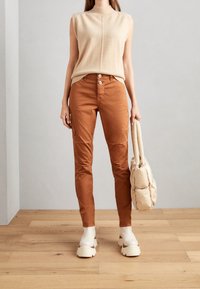 Beige bezrukávový pletený top, rustově zbarvené přiléhavé kalhoty, robustní béžové dřeváky a krémová nadýchaná kabelka. Pozadí tvoří dřevěná podlaha.