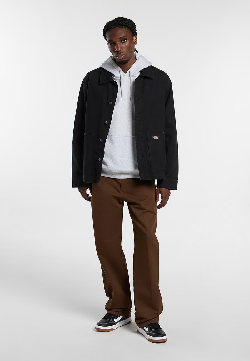 Giacca nera con colletto classico, chiusura frontale con bottoni e due tasche. Indossata sopra una felpa con cappuccio grigio chiaro, pantaloni marroni e scarpe da ginnastica nere.