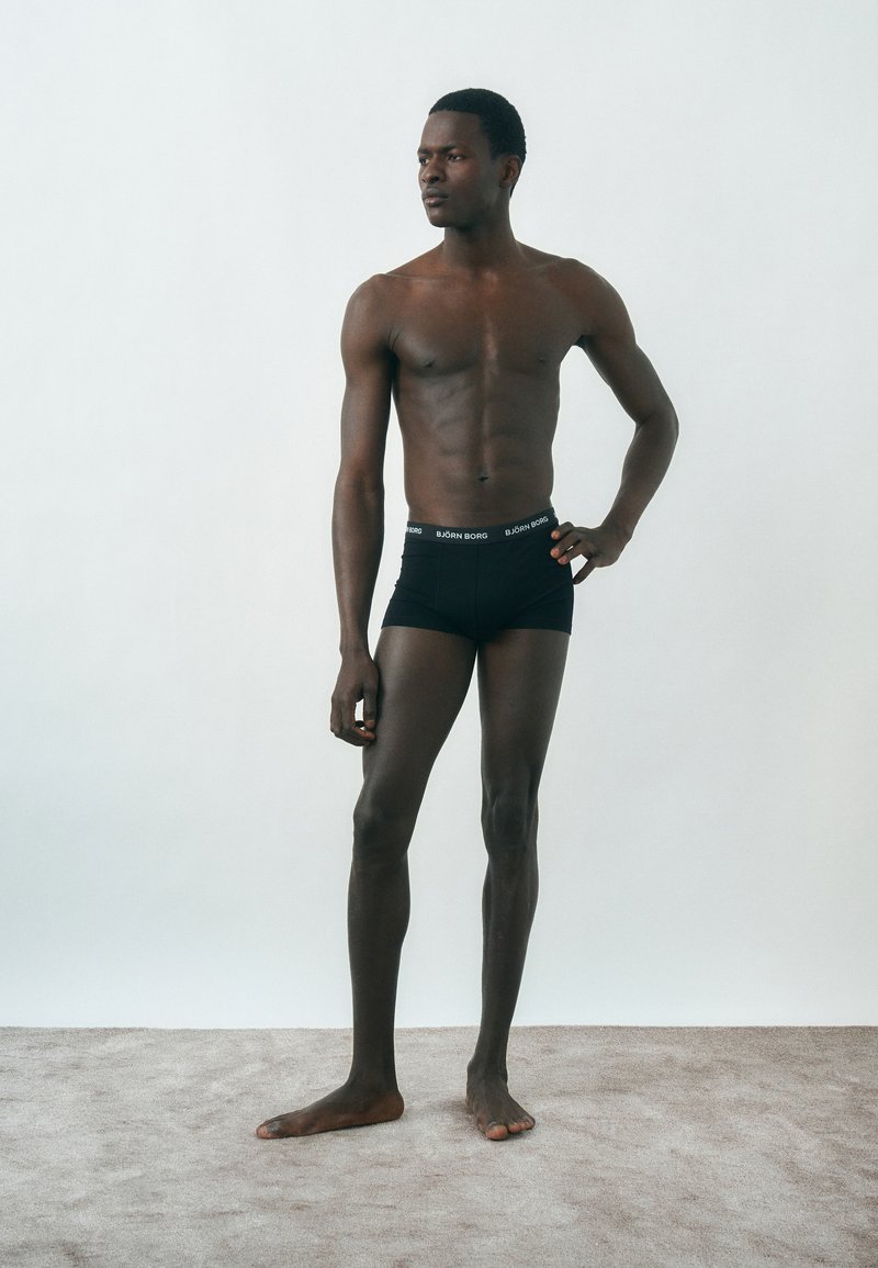 Αθλητικός άντρας στέκεται ξυπόλυτος σε χαλί, φοράει μαύρα μποξεράκια Björn Borg και κοιτάζει στο πλάι με το ένα χέρι στη μέση.