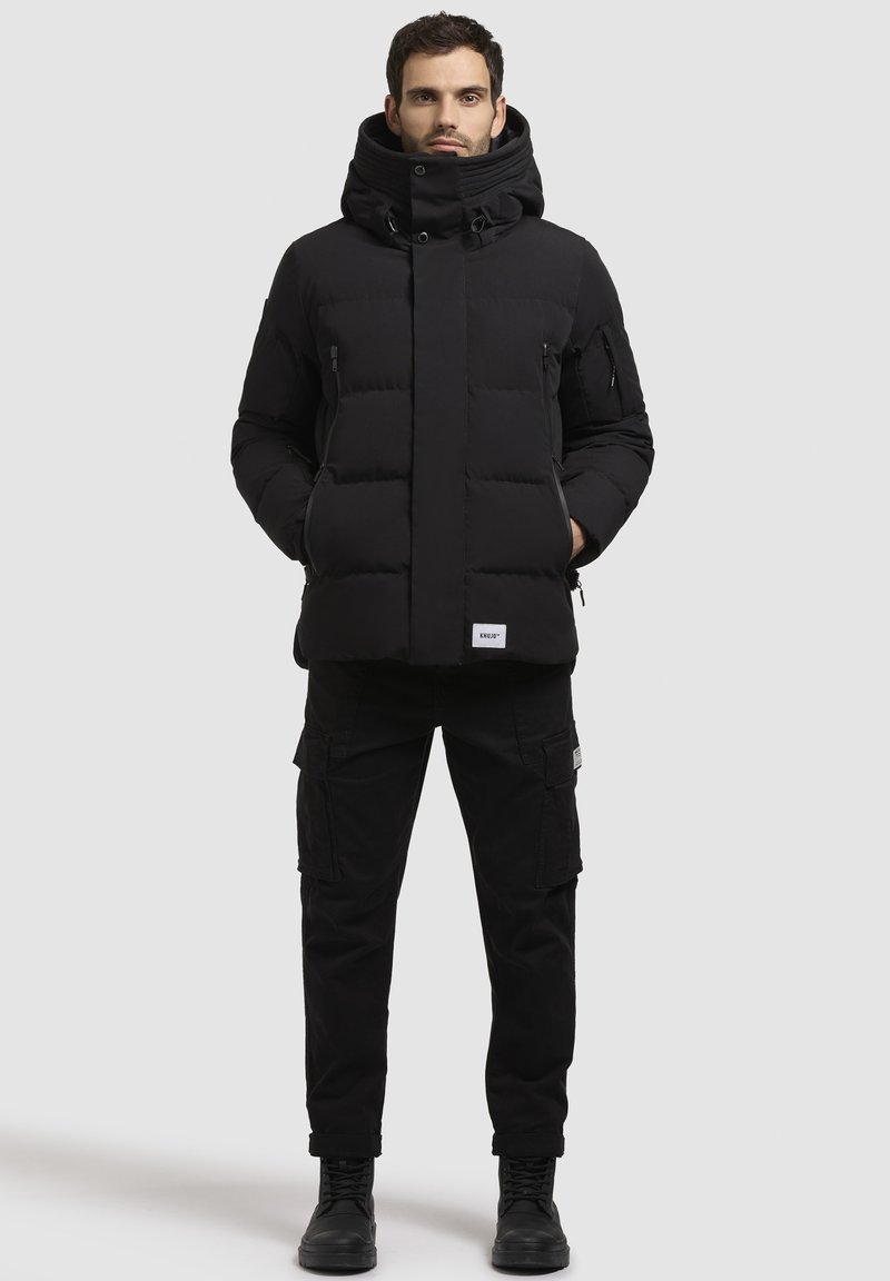 Jachetă pufită neagră cu guler înalt și glugă, având secțiuni căptușite și fermoare laterale. Pură cu pantaloni cargo negri și cizme.