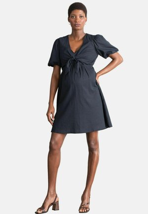 Schwarz gestreiftes Kleid mit geknotetem Vorderteil, kurzen Puffärmeln und tailliertem Oberteil, das in einen knielangen Rock übergeht. Kombiniert mit dunklen Sandalen.