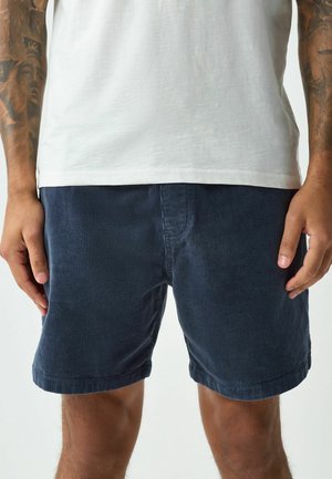 Mann trägt dunkelblaue Cord-Shorts und ein schlichtes weißes T-Shirt, steht mit entspannten Händen seitlich am Körper.