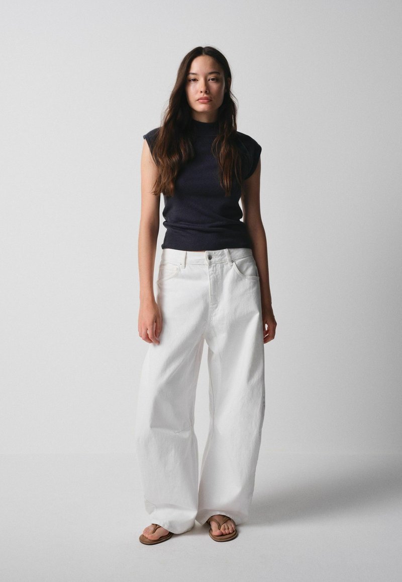 Woman with long dark hair wearing a sleeveless black top, wide white pants, and flip-flops standing against a plain light background.