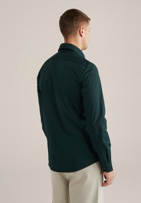 Donkergroene overhemd met lange mouwen met een klassieke kraag, knoopmanchetten en een gladde katoentextuur, gedragen over lichtgekleurde broek.