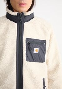 Jachetă din fleece crem cu guler înalt, având un buzunar pe piept cu fermoar gri. Material texturat cu un accent de fermoar neted și închis la culoare.
