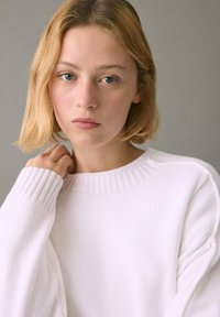 Beli pleteni pulover z rebrastim izrezom in manšetami; mehka tekstura; model rahlo dotika vratu, nevtralno ozadje.