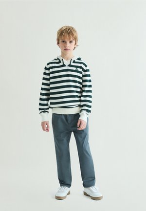 Boy with blond hair wearing a white and dark green striped hoodie, gray pants, and white sneakers standing against a plain background.