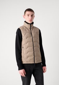 Beige dunjakke med høy krage, glidelås og quiltemønster; kombinert med en svart genser med lange ermer og mørke jeans.