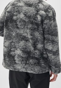 Osoba w szarej i czarnej strukturze polarowej kurtce i czarnych spodniach, widoczna z tyłu na jednolitym tle.