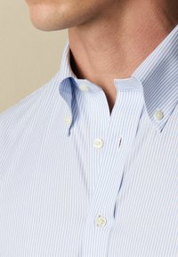 Hellblaues Button-Down-Shirt mit weißen vertikalen Streifen, ausgestattet mit einem Kragen, zwei Knopfakzenten und einer strukturierten Stoffoberfläche.