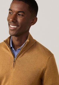 Sweat à zip marron en tissu doux, avec un col montant et superposé avec une chemise rayée bleu et blanc en dessous.
