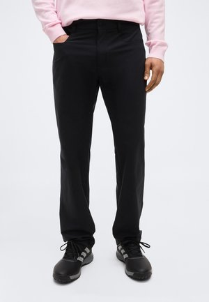 Mees, kes kannab musti pükse, musti tenniseid hallide triipudega ning heleroosat pika varrukaga särki, seisab ühe käega taskus.