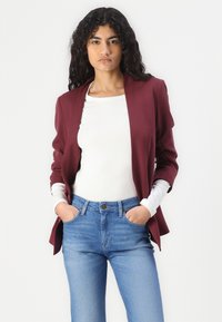 Bordový blazer vyrobený z lehkého materiálu s otevřeným předním designem, kombinovaný s bílým topem a světlými modrými džíny. Ruce umístěné podél těla.
