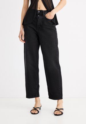 Személy fekete, laza, magas derekú farmert visel, egyik kezét a zsebben tartja, fekete sarokszandállal és áttetsző fekete felsővel párosítva.