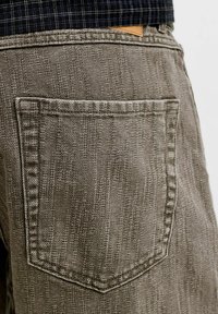 Gros plan de la poche arrière texturée grise d’un pantalon portée avec une chemise foncée à motifs rentrée.