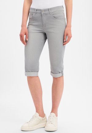 Woman wearing light gray rolled-up denim capri pants and white lace-up sneakers, standing against a plain white background.