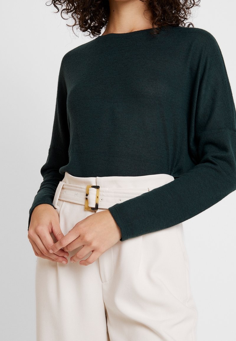 Femme portant un haut à manches longues vert foncé rentré dans un pantalon blanc taille haute avec une large ceinture blanche et une boucle rectangulaire.