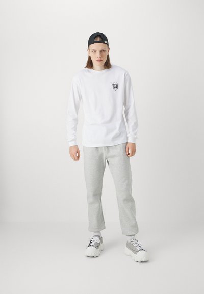 Long-sleeve white shirt with a black logo on the chest, paired with light grey sweatpants and chunky white and grey sneakers, styled with a black cap.