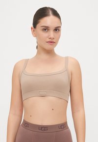 Bežasti rebrasti bralet s uskim trakama i remenom s logom. Model nosi smeđe tajice visokog struka. Glatka tekstura, minimalni dizajn.