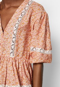 Robe rose à imprimé marguerites blanches, avec bordure en dentelle blanche sur un col en V profond, aux manches et à la taille froncée, portée par une personne à la peau brune.