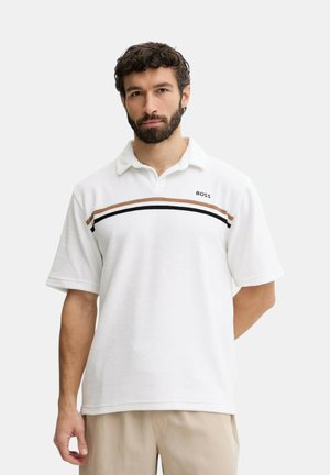 Uomo con barba che indossa una polo bianca con strisce marroni e nere sul petto e pantaloni beige, in piedi davanti a uno sfondo semplice.