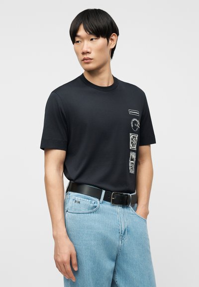 Νέος άνδρας φοράει μαύρο γραφικό t-shirt μπλουζάκι μέσα σε ανοιχτόχρωμο μπλε τζιν με μαύρη ζώνη, κοιτάζοντας πλάγια σε μονόχρωμο φόντο.