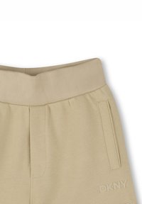 Pantalones de chándal beige con una cinturilla acanalada, bolsillos laterales y el logo de DKNY grabado en la pierna izquierda. Textura de tejido suave en todo el conjunto.