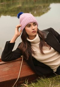 Roosa kudumüts sinise pom-pomiga, kombineeritud kreemja kaelusega ja musta manteliga, looduse taustal.