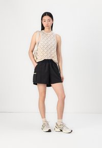 Top senza maniche beige in maglia con motivo a diamante; pantaloni corti neri con tasca laterale; scarpe da ginnastica beige e grigie; sfondo neutro.