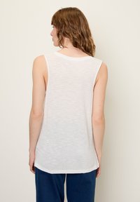 Balta be rankovių palaidinė, pagaminta iš lengvos, šiek tiek tekstūruotos medžiagos. Apvalus iškirpimas ir kreivė į apačią. Modelis vilki mėlynas kelnes.