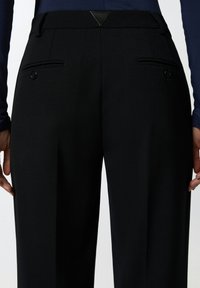 Crne krojene hlače s glatkom teksturom, dva stražnja džepa s gumbima i trokutastim kožnim detaljem na pojasu.