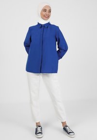 Camisa azul de botón con cuello, combinada con pantalones blancos y zapatillas de deporte azul marino. La camisa tiene mangas largas y un corte relajado.