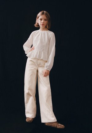 Młoda osoba z krótkimi włosami, ubrana w białą bluzkę z długim rękawem, szerokie kremowe spodnie z haftem oraz brązowe mokasyny, stojąca na tle czerni.