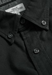 Camicia nera con bottone, realizzata in un tessuto testurizzato, con colletto classico e bottoni neri. L'etichetta riporta "next, crafted for the individual."