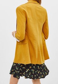 Gul kavaj med texturerad tyg, snibbkrage och två sidofickor, bärs över en svart volangkjol med blommönstrade accenter.