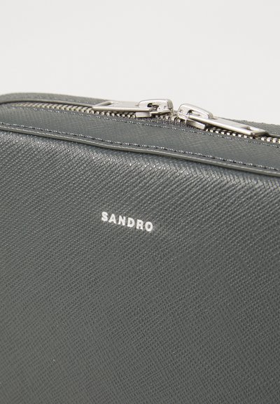 Pochette in pelle grigia con texture, dotata di doppia zip argentata e nome del marchio "SANDRO" impresso in lettere bianche.