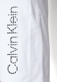 Tissu blanc avec logo Calvin Klein noir vertical imprimé le long de la couture d’un vêtement, probablement une manche ou une jambe de pantalon.
