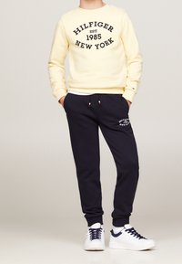 Kind trägt einen cremefarbenen Sweatshirt mit dem Aufdruck "Hilfiger Est. 1985 New York", marineblaue Jogginghosen und weiße Sneakers und steht mit den Händen in den Taschen.