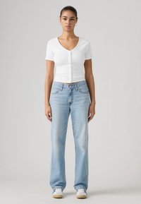 Γυναίκα που στέκεται με το πρόσωπο προς τα εμπρός, φορώντας λευκό κοντομάνικο πουκάμισο με κουμπιά, ανοιχτό μπλε ίσια τζιν παντελόνια, σκουλαρίκια κρίκους και λευκά αθλητικά παπούτσια.