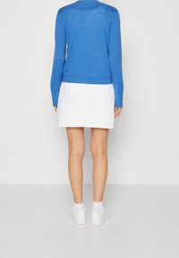 Blauer Pullover mit langen Ärmeln und strukturiertem Design, kombiniert mit einem weißen Rock. Ein schlichter, moderner Look mit glatten Stoffen. Weiße Sneakers abgebildet.