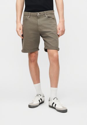 Mężczyzna w podwiniętych oliwkowozielonych szortach, białych skarpetach za kostkę i białych tenisówkach z czarnymi paskami, stojący na tle jednolitego białego tła.