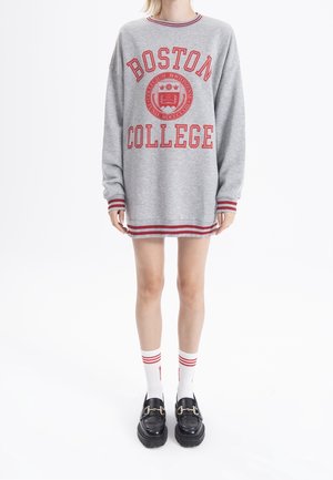 Sweatshirt gris avec le texte et l'emblème "BOSTON COLLEGE" en rouge. Comprend des poignets et un ourlet rayés. Porté avec des chaussettes blanches et des chaussures à plateforme noires.