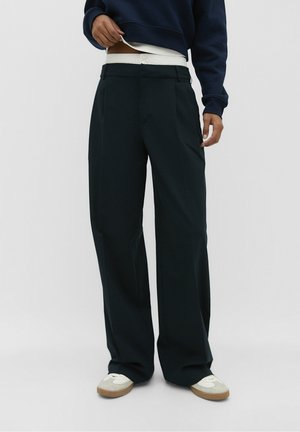 Personne portant un pantalon noir ample, des baskets blanches et un sweat-shirt bleu marine, soulevant le sweat-shirt pour révéler une chemise blanche en dessous.