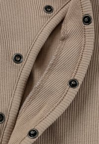 Beige ribstof met zwarte drukknoopjes langs de rand, met nette stiksels en een gladde textuur.