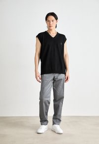Svart ärmlös stickad topp med djup V-ringning och texturerad mönster, kombinerad med ljusgrå byxor och vita sneakers.