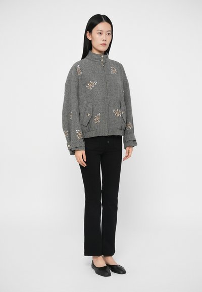 Sieviete, kura valkā pelēku džemperi ar ziedu izšuvumiem, melnas pieguļošas bikses un melnas plakandobes, stāv uz vienkārša fona.